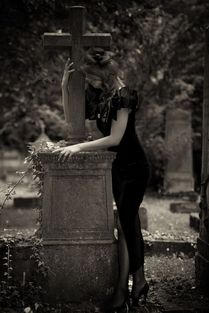 Beauty & Fashion Aufnahme - junge Frau in schwarzem Kleid und mit Tüll bedecktem Gesicht trauert an einem Grabkreuz