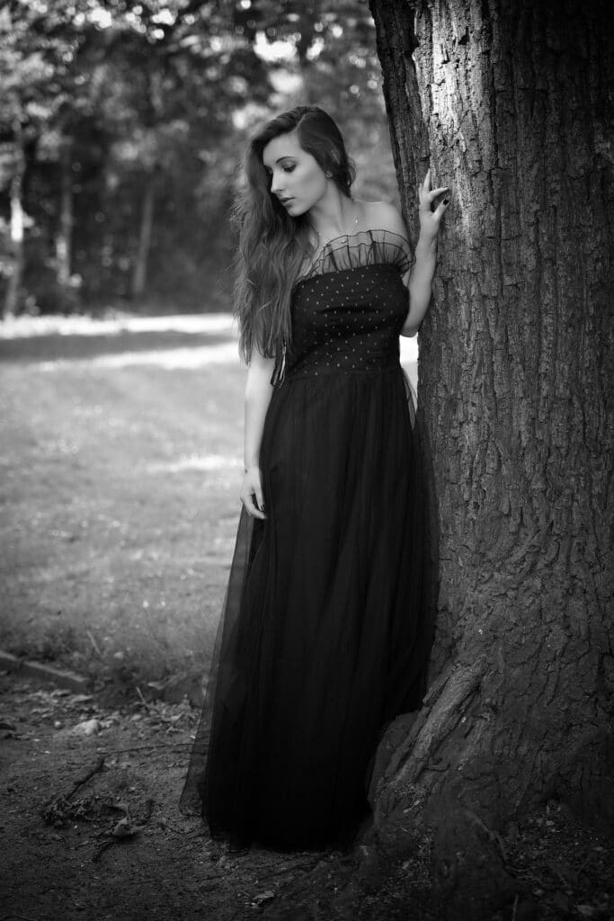 Beauty & Fashion Aufnahme - junge Frau steht in langem schwarzen Kleid und hält sich an einem Baum fest
