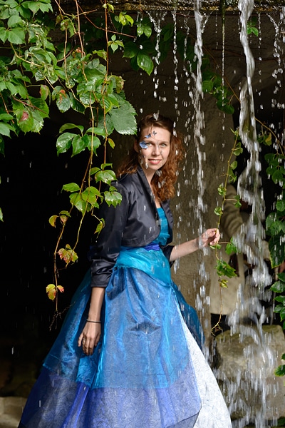 Elfia - stehende junge Frau in blauem Kleid hinter einem Wasserfall