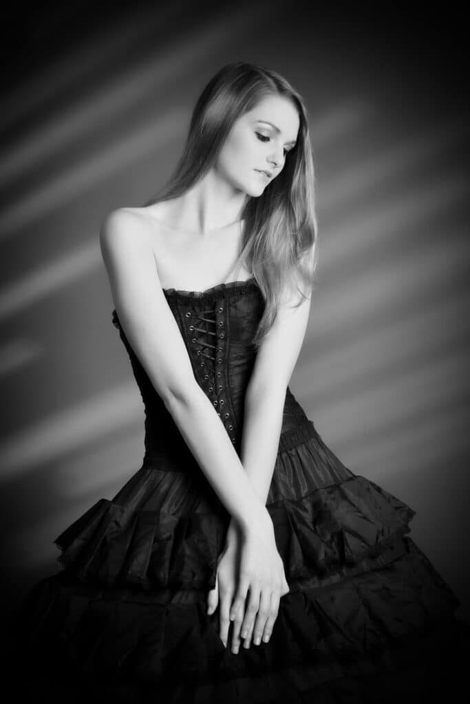 Beauty & Fashion Aufnahme - junge Frau in schwarzer Korsage und langem gestuften schwarzen Rock steht vor gestreiftem Hintergrund
